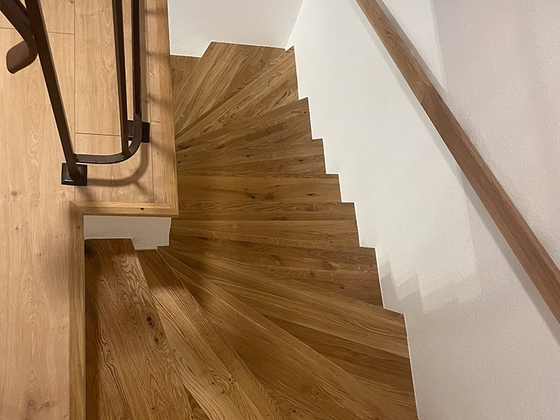 Treppenrenovierung planen: Aufmaß, Anschlüsse und Ablauf 1 Blick auf einen gewendelten Treppenlauf mit holzfarbener Oberfläche und dunklem Handlauf